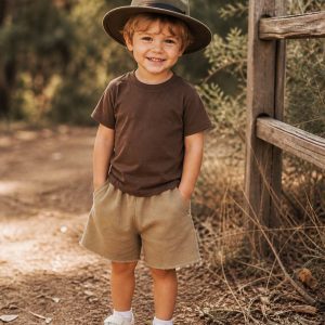 boy beside a wooden fence with a brown t-shirt, long fawm shorts and an Akruba style hat