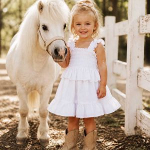 little girl wearing a kids on trend white frilly dress standing by a white pony